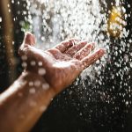 A man’s hand in a spray of water in the sunlight against a dark background. Water as a symbol of purity and life